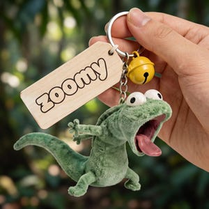 May include: A keychain featuring a plush green lizard with wide eyes and an open mouth with a pink tongue, attached to a silver metal ring with a small yellow bell and a light-colored wooden rectangular tag engraved with the word ZOOMY.