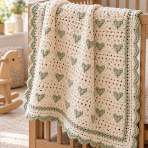 May include: A cream-colored crocheted blanket featuring a repeating pattern of small sage green hearts. The blanket has a scalloped sage green border and is draped over the side of a wooden crib, showcasing the intricate yarn texture and design.