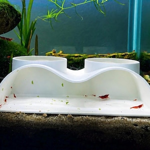 May include: A white plastic aquarium feeding station featuring two circular openings on top and a wide, curved front platform. Several small, bright red cherry shrimp are positioned on the flat surface of the feeder inside the glass tank.