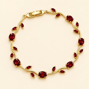 May include: A gold-tone bracelet featuring a vine-like design with alternating oval-shaped red gemstones and small leaf-shaped red accents. The bracelet is secured with a rectangular box clasp mechanism, creating a continuous circular link structure.