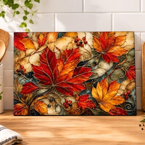 May include: A rectangular glass cutting board featuring a vibrant autumn-themed design with detailed red, orange, and yellow maple leaves, dark berries, and swirling vine patterns set against a textured, multi-colored background.