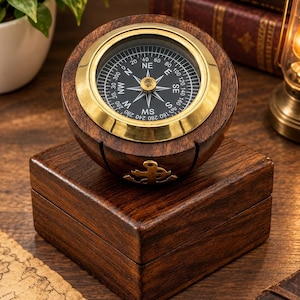 May include: A round wooden compass with a brass rim and a black dial featuring white cardinal directions and degree markings. The compass is mounted on a square wooden base, which displays a small brass anchor emblem on its front side.