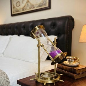 May include: A brass hourglass with a rotating frame and purple sand, mounted on a circular base shaped like a ship's wheel. The hourglass is tilted at an angle within its support structure, featuring clear glass bulbs and a metallic finish.
