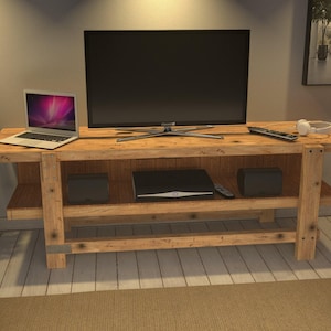 May include: A rectangular wooden media console featuring a natural light brown finish with visible wood grain and knots. The unit has a flat top surface and a lower shelf, supported by four thick, sturdy legs. It holds a television, a laptop, and speakers.