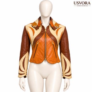 Pode incluir: Um casaco curto de couro da Usvora Industries com um design de blocos de cores em tons de castanho, laranja e creme. O casaco possui um fecho central dourado, dois bolsos com fecho, uma gola pontiaguda e detalhes em painéis curvos.