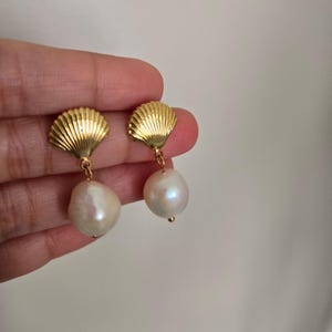Peut inclure: Une paire de boucles d'oreilles dorées composées d'une partie supérieure en forme de coquillage texturé en éventail, attachée à une perle blanche irrégulière. Les éléments métalliques sont polis et les perles présentent une surface lisse et nacrée.