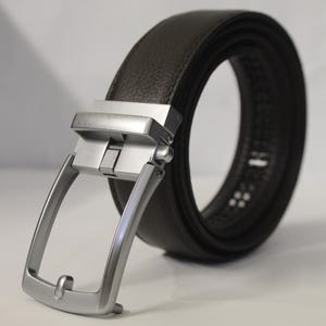 May include: A dark brown textured belt featuring a silver-toned metal buckle with a rectangular frame and a sliding ratchet mechanism. The belt is coiled into a circular shape, showcasing the leather-like material and the metallic hardware finish.