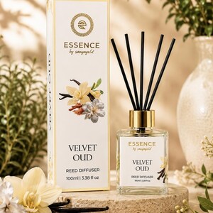 Op de afbeelding: Een vierkante glazen geurstokjesdiffuser met een gouden dop en vijf zwarte stokjes, vergezeld door een rechthoekige doos. Beide bevatten de tekst Essence by sanyagold, Velvet Oud, 100ml en zijn versierd met illustraties van vanillebloemen.