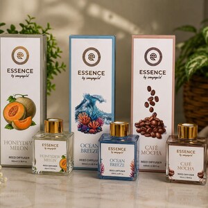 May include: Three rectangular reed diffuser glass bottles with gold caps and matching tall boxes labeled Essence by sanyagold. The scents are Honeydew Melon with a green box, Ocean Breeze with a blue box, and Café Mocha with a brown box, each 100ml.