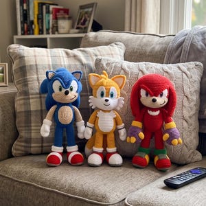 May include: Three handmade crochet plush toys representing Sonic the Hedgehog, Tails, and Knuckles. Sonic is blue with white accents, Tails is yellow and white with two tails, and Knuckles is red with green and purple details on the hands.