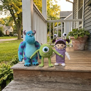 May include: Three handmade crochet dolls representing characters from Monsters Inc. A large blue fuzzy-textured monster with purple spots, a small round green one-eyed monster, and a human child wearing a purple monster costume with two eyes.
