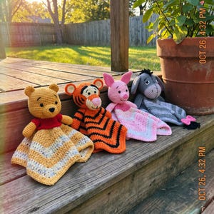 May include: Four crochet lovey blankets featuring stuffed animal heads: a yellow bear with a red shirt, an orange and black striped tiger, a pink pig, and a grey donkey. Each toy has a soft, textured blanket body with chevron patterns.