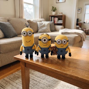 Könnte beinhalten: Drei gehäkelte Minion-Puppen in unterschiedlichen Höhen, jeweils mit gelben Körpern, blauen Latzhosen, schwarzen Schuhen und silberfarbenen Brillen. Die größte Puppe hat dünnes schwarzes Haar, während die kleinste einen kleinen braunen Teddybär hält.