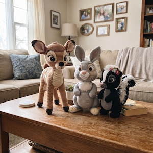 May include: Three crocheted stuffed animals featuring a brown deer with white spots, a light grey rabbit with a white chest, and a black and white skunk with a floral crown on its head. The figures are crafted with detailed yarn stitching.