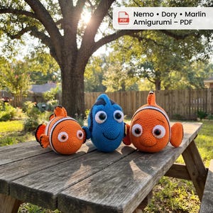 May include: Three crocheted amigurumi fish toys featuring Nemo, Dory, and Marlin characters. The toys are orange, white, and blue with large plastic eyes. A digital label at the top reads: Nemo • Dory • Marlin Amigurumi – PDF.