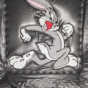 May include: A black garment featuring a square, glossy, black patch with an embroidered graphic of the cartoon character Bugs Bunny. The character is depicted in a running pose with grey fur, pink inner ears, and a red mouth, stitched onto the fabric.