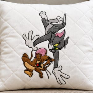 May include: A white quilted throw pillow featuring a central embroidered graphic of the cartoon characters Tom and Jerry. The grey cat Tom is reaching down toward the brown mouse Jerry, who is positioned below him. The embroidery uses black, grey, brown, pink, and white thread to depict the characters in a dynamic pose.