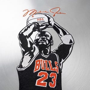 May include: An embroidered patch featuring a basketball player in a black and orange Bulls jersey with the number 23, holding a basketball above their head. The design includes the text NBA on the ball and a signature in orange thread above the player.