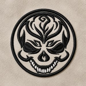 Puede incluir: Un parche bordado circular con un diseño de calavera en blanco y negro. La calavera está estilizada con intrincadas líneas negras que forman las cuencas de los ojos, la nariz y los patrones de la frente, con dientes en hilo blanco.