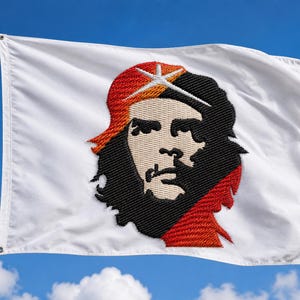 Op de afbeelding: Een witte rechthoekige vlag bevestigd aan een zilveren metalen mast, met een groot, centraal geborduurd portret van Che Guevara. Het portret gebruikt zwarte, beige en rode draad om het gezicht en de baret op de effen witte stof weer te geven.