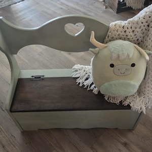 Puede incluir: Un banco de madera verde salvia con un recorte en forma de corazón en el respaldo y un asiento abatible de color marrón oscuro. Un peluche de vaca highland verde claro y una manta de ganchillo color crema con flecos descansan sobre el banco.