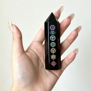 Puede incluir: Una torre de cristal de obsidiana negra pulida con forma de prisma hexagonal y punta superior. La cara frontal presenta siete símbolos de chakra alineados verticalmente, cada uno representado en un color distinto que incluye morado, azul, verde, amarillo y rojo.