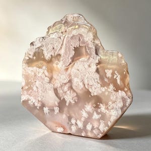 Puede incluir: Una rodaja de ágata flor pulida y semitranslúcida con tonos rosa suave y melocotón. La piedra muestra intrincadas inclusiones florales blancas en forma de pluma dispersas por su superficie, creando un patrón natural y orgánico.