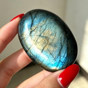 Pode incluir: Uma pedra preciosa de labradorite de formato oval com uma superfície polida e lisa, apresentando reflexos iridescentes em azul e dourado. A pedra exibe fraturas internas naturais e tons de cinza escuro por toda a sua estrutura.