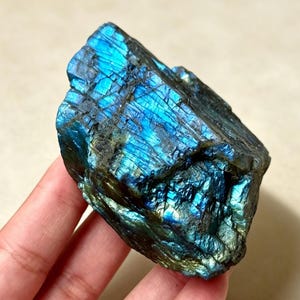 Peut inclure: Un spécimen minéral de labradorite brute présentant une surface sombre et irrégulière avec des reflets irisés bleus et dorés. La pierre affiche une texture naturelle non polie avec des fissures profondes et des couches cristallines.