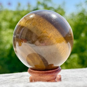 Puede incluir: Una esfera de piedra ojo de tigre pulida con bandas de fibras chatoyantes de color marrón dorado y marrón oscuro. El orbe liso y reflectante se muestra descansando sobre un pequeño soporte de madera tallada con base redondeada.