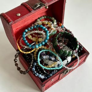 Puede incluir: Un cofre de madera roja con un cierre de metal, lleno de una colección de pulseras de cuentas variadas. Las pulseras presentan diversos colores, incluyendo cuentas azules, verdes, amarillas, negras y transparentes, con texturas facetadas o lisas.