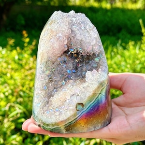 Puede incluir: Una geoda de amatista vertical con un revestimiento metálico iridiscente y pulido en la base exterior. El interior presenta puntas cristalinas de color violeta claro que reflejan colores del arcoíris, creando un efecto brillante sobre la superficie dentada.
