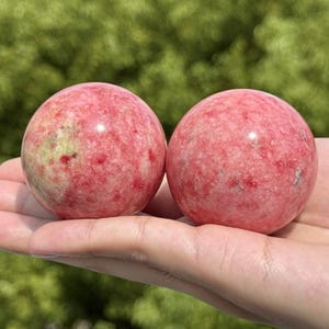 Op de afbeelding: Twee gepolijste bolvormige stenen met een gevlekte textuur in tinten roze, rood en zachtgroen. De oppervlakken zijn glad en reflecterend, wat de natuurlijke minerale variaties in deze twee ronde stenen bollen duidelijk zichtbaar maakt.