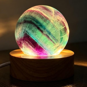 May include: A spherical fluorite crystal with bands of purple, green, and white, illuminated from beneath by a circular wooden base. The stone features natural internal fractures and colour variations, resting on a light-coloured wood stand.