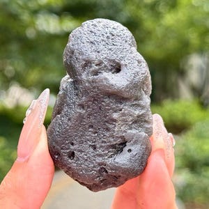 Puede incluir: Un espécimen mineral gris oscuro translúcido con una textura de superficie rugosa, picada e irregular. La piedra presenta una forma redondeada y bulbosa con pequeñas hendiduras y detalles porosos en todo el exterior, mostrando un aspecto natural y bruto.