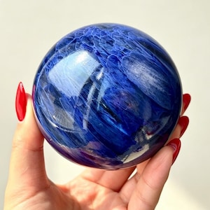Peut inclure: Une pierre sphérique polie présentant des teintes bleu indigo et bleu roi avec des motifs fibreux tourbillonnants et des inclusions réfléchissant la lumière sur toute la surface. L'objet lisse et arrondi affiche une finition brillante.