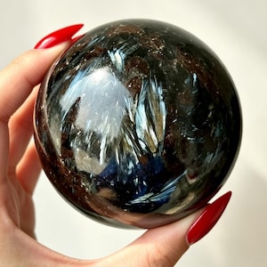 Puede incluir: Una piedra esférica pulida que presenta un color base marrón oscuro y negro profundo con prominentes inclusiones fibrosas de color azul iridiscente y plateado que crean un efecto reflectante y chatoyante en la superficie lisa y redondeada.