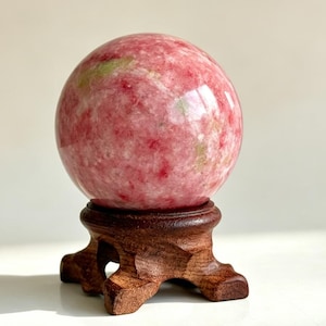 Pode incluir: Uma pedra esférica polida com um padrão manchado de rosa vibrante, branco suave e inclusões minerais verde-pálidas. A esfera repousa firmemente sobre um suporte de tripé de madeira esculpida em castanho escuro com uma base lisa e arredondada.