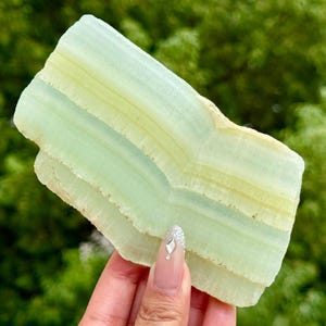 Puede incluir: Una losa plana y rectangular de mineral verde pálido con bandas horizontales distintivas y una apariencia translúcida y estratificada. La piedra presenta variaciones sutiles en tonos verde claro y blanquecino, con un borde rugoso y texturizado.
