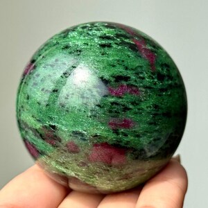 Puede incluir: Una piedra esférica pulida que presenta un color base verde vibrante con inclusiones prominentes de depósitos minerales de color magenta intenso y negro en toda la superficie, creando una textura veteada natural en la forma.