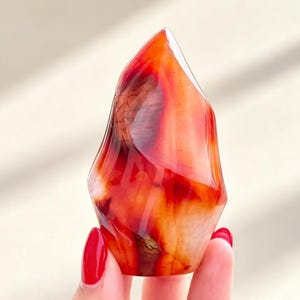 May include: A polished carnelian crystal flame carving featuring vibrant gradients of deep red, burnt orange, and translucent amber tones. The stone has a smooth, glossy surface with natural internal banding and a pointed, flame-like silhouette.