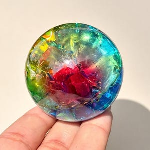 May include: A spherical paperweight made of clear resin containing a vibrant array of multicoloured inclusions. The interior features fragments of red, blue, green, and yellow foil that create a prismatic effect throughout the transparent orb.