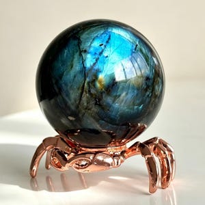 May include: A polished labradorite sphere displaying iridescent blue and gold flashes across its dark, mottled surface. The sphere rests securely on a metallic rose gold-coloured stand shaped like a crab with articulated legs and claws.
