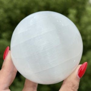 Op de afbeelding: Een gladde, bolvormige selenietkristal met een doorschijnend wit uiterlijk en een vezelige, gelaagde textuur. Het oppervlak van de steen reflecteert het licht en toont subtiele horizontale banden en een gepolijste, ronde vorm over het hele object.