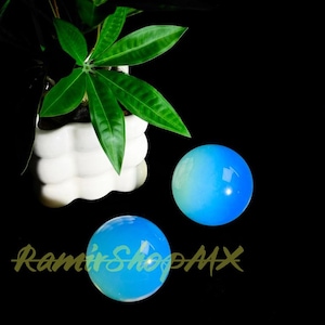 Op de afbeelding: Twee gladde, bolvormige blauwe stenen met een doorschijnend, gloeiend uiterlijk. De bollen zijn geplaatst bij een witte, getextureerde keramische plantenbak met een groene plant met puntige bladeren. De tekst RamirShopMX is zichtbaar.