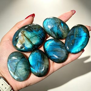 May include: Six polished, oval-shaped labradorite stones with a smooth texture. Each stone displays a vibrant, iridescent blue and teal flash across its surface, with visible natural internal fractures and dark grey to black base tones.