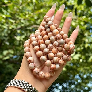 Puede incluir: Una pulsera de cuentas compuesta por piedras redondas y pulidas con un aspecto jaspeado en tonos crema, beige y marrón rojizo. Las cuentas están ensartadas en un bucle continuo, mostrando una textura suave y brillante en toda la pieza.
