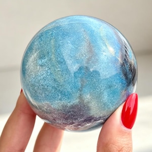 Puede incluir: Una piedra esférica pulida que presenta una mezcla vibrante de tonos azul translúcido y morado intenso. La superficie muestra patrones en espiral con sutiles inclusiones internas y reflejos de luz en toda la forma mineral redondeada.