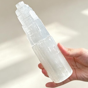 Può includere: Una torre di cristallo di selenite bianca traslucida con una struttura cilindrica a gradini. Il minerale presenta una consistenza fibrosa e stratificata con una finitura lucida, che si restringe verso l'alto in una serie di segmenti circolari impilati.