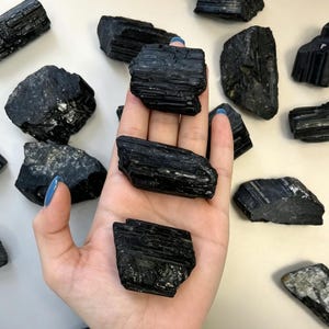 Può includere: Una collezione di pietre di tormalina nera grezza con strutture cristalline colonnari striate e bordi irregolari. Le pietre sono opache, di colore nero intenso e presentano una consistenza naturale non levigata con solchi.